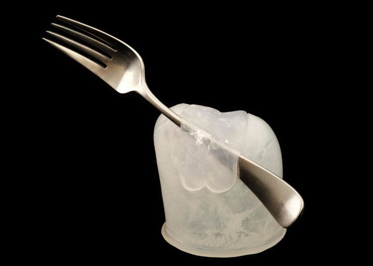 Studio shot of a silver fork situated in an white opaque molded cap that fits precisely on Cindy's residual right hand. The fork is propped against the silicone and a layer of silicone wraps over the fork to hold it in place at the angle that best mimics the angle of a fork in a hand.