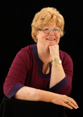 Headshot of Cindy smiling, wearing right cosmetic hand, with her right arm resting on table in front of her and her chin propped up by her left hand.