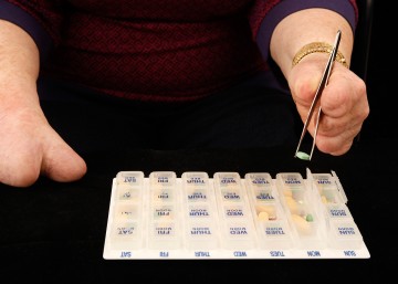 Cindy using a pair of all-silver tweezers to select pills from a clear plastic pillbox. The pillbox has days and times printed on small individual boxes, and a few of the boxes are open and showing pills of assorted sizes and colors (yellow, red, green). Cindy is using the tweezers with her left hand, and her torso is visible in the background. Her right hand is visible to the side.