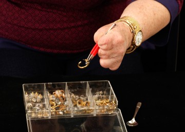 Cindy using the red-cushioned tweezers to select gold an earring from the clear plastic earring storage box. Cindy is using the tweezers with her left hand, and it holds one gold hoop earring. Her torso is visible in the background, and the demitasse spoon is on the side.