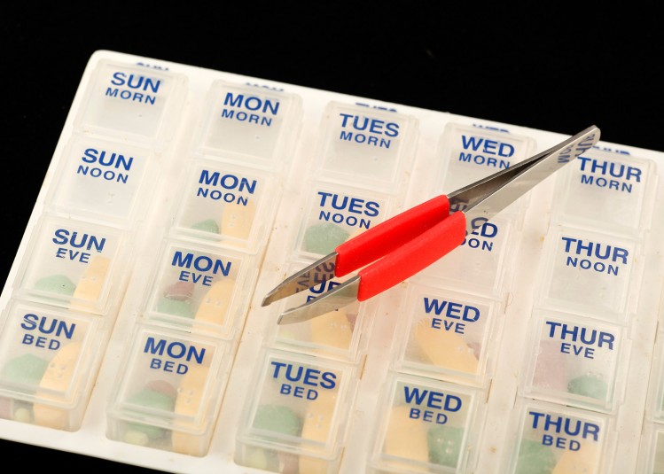 Close-up of the clear-plastic pillbox with a tweezers lying on top and all pillbox doors closed. The tweezers have a red soft grip.