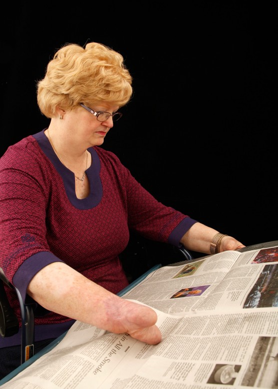 Cindy props the newspaper board on her lap. Its surfaces and structure allow her to view and page through a full size two page spread of a standard newspaper.