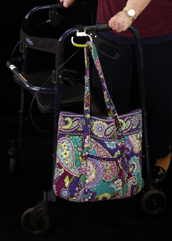 Studio close up of carabiner attached to the left handle of the walker, and angled down. It holds a paisley pastel-colored quilted cloth bag. Cindy'd hands are visible, holding the walker handles.