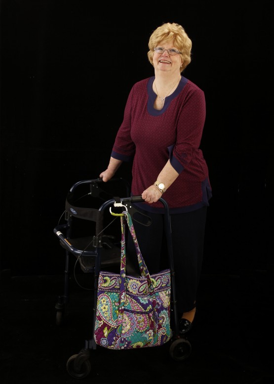 Studio shot of Cindy standing at her walker against a black background. Cindy is in a maroon shirt, is smiling at the camera, and has her two hands on the handles of the walker. The carabiner is attached to the left handle of the walker, and is angled down. It holds a paisley pastel-colored quilted cloth bag.
