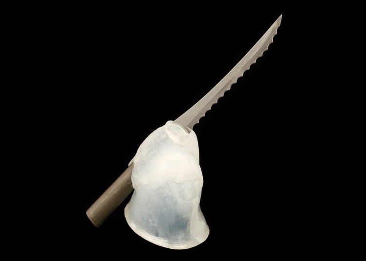 Studio shot of a brown plastic-handled metal serrated dinner knife situated in an white opaque molded cap that fits precisely on Cindy's residual right hand. The cap sits upright and the knife angles from bottom left to upper right of the screen. The knife is propped against the silicone and a layer of silicone wraps over the fork to hold it in place at the angle that best mimics the angle of a knife in a hand.