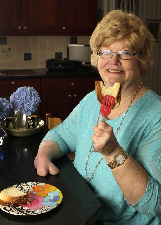 Cindy using the salad tongs. She is in her kitchen and smiling at the camera, wearing a turquoise shirt. Her left hand is holding the tongs, to show us how they grip the sandwich half. The sandwich, on white bread, has one bite out of it. The other half of the sandwich is on a flowered plate on the table in front of Cindy.