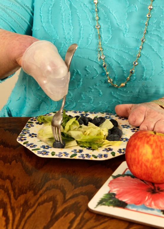 Shot of Cindy's sitting at a brown dining table, using the silicone fork unit. Cindy is wearing a turquoise shirt and gold necklace, and we see her body from just below the neck to where it is hidden behind the table where she is sitting.  The fork is in her right hand and she is piercing a blueberry with it. The plate of food includes lettuce and blueberries, and there is a nearby cutting board with an apple on it.