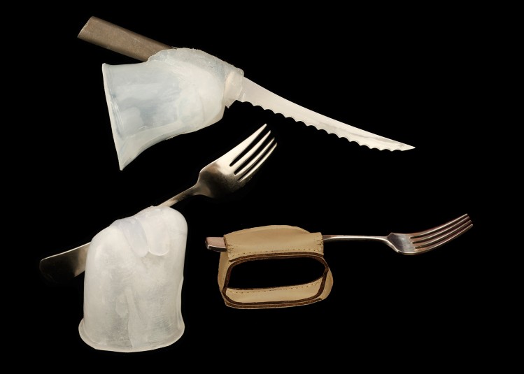 Studio shot of the fork/silicone and knife/silicone units, as well as the fork in the leather cuff. The knife is at the top left of the screen, angled from top left to bottom right; the fork is at the bottom left of the screen; the fork in the cuff is at bottom right.